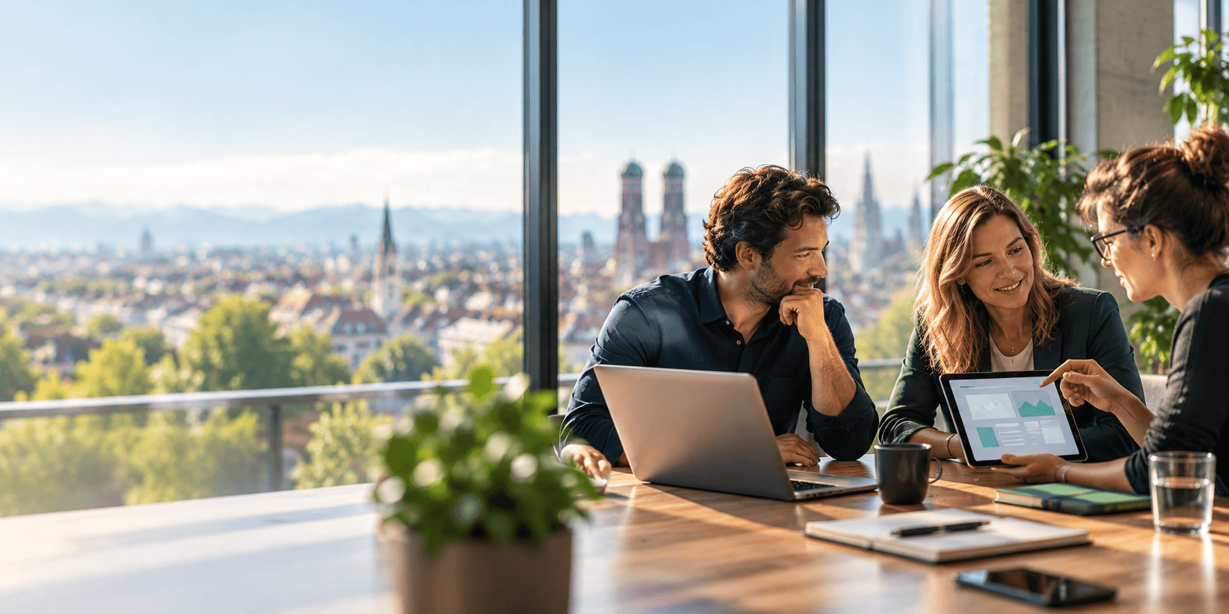 Beratungsgespräch an einem Tisch mit Laptop und Tablet, im Hintergrund die Münchner Frauenkirche und die Alpen